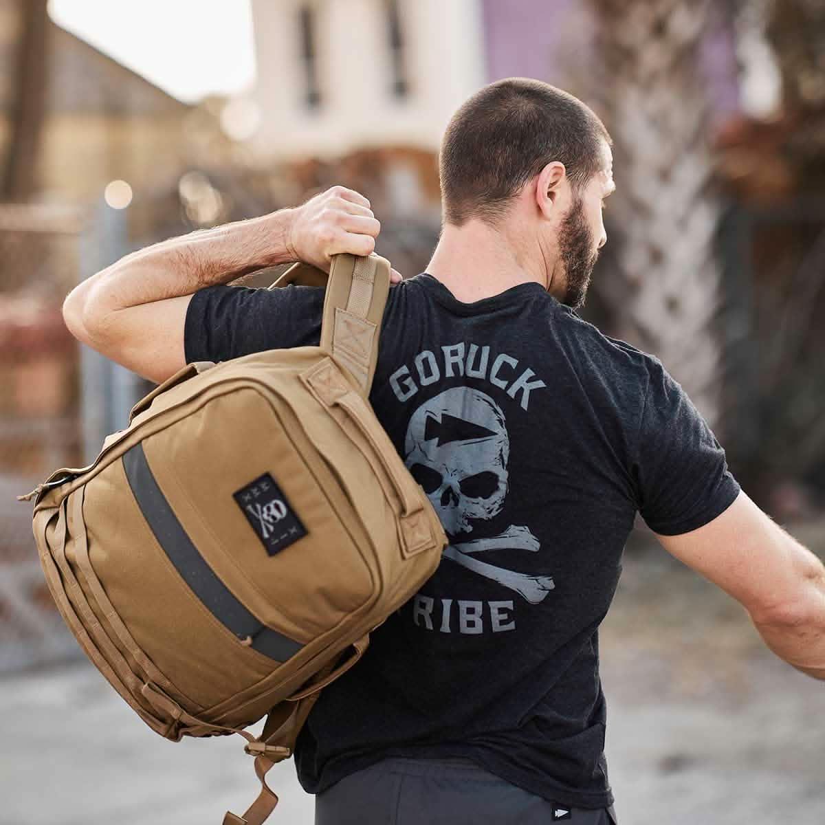 Man wearing GORUCK Tribe t-shirt carrying tan rucksack outdoors, rugged fitness gear